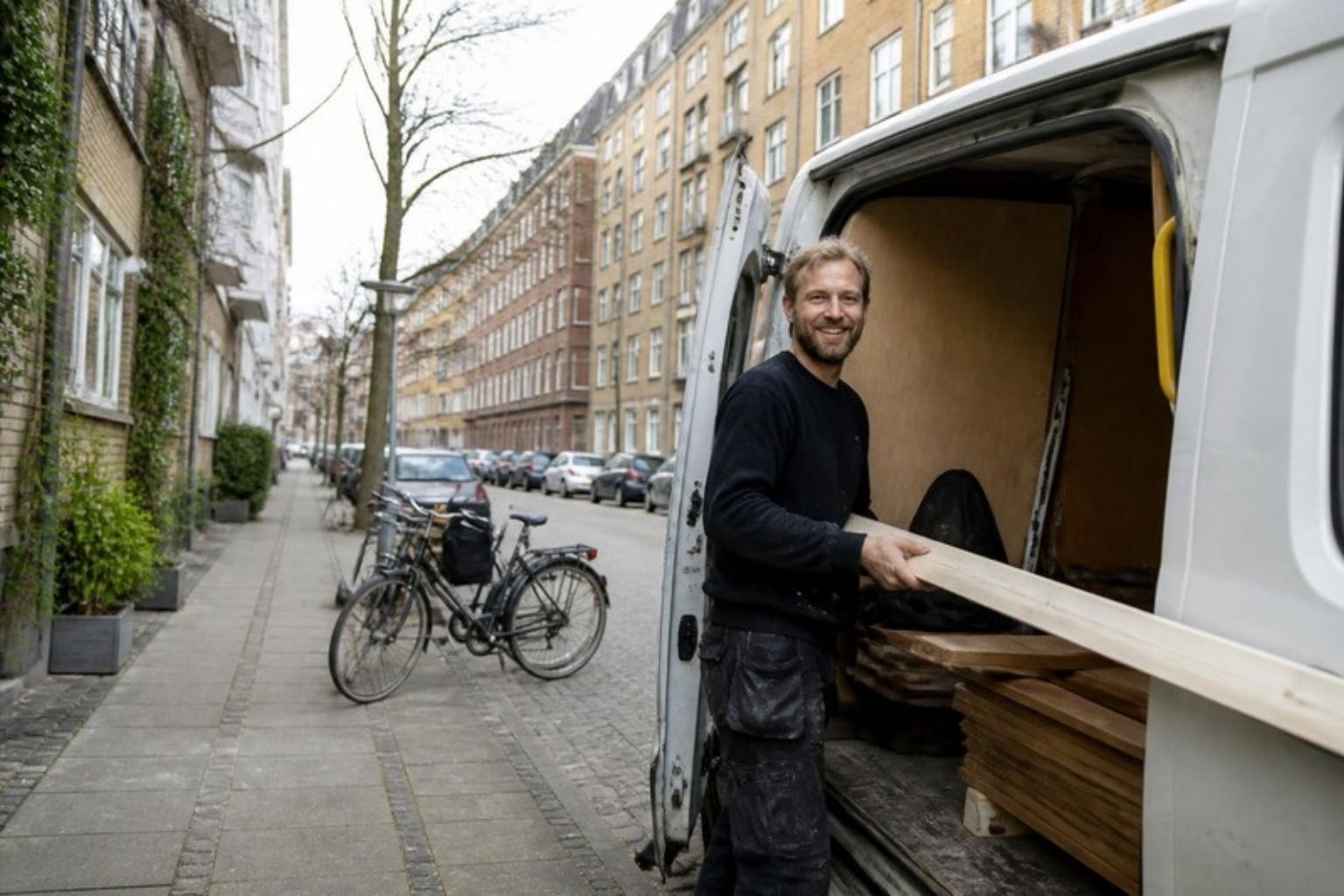 Smilende tømrer bærer værktøj fra en varevogn på en gade i København